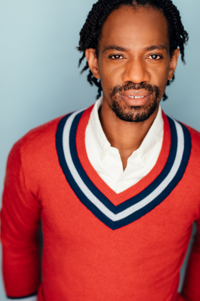 Audition Headshot for Reggie White. Reggie looks into the camera in front of a light blue background while wearing a red sweater with a blue and white stripe along the outline of the neck cutout.